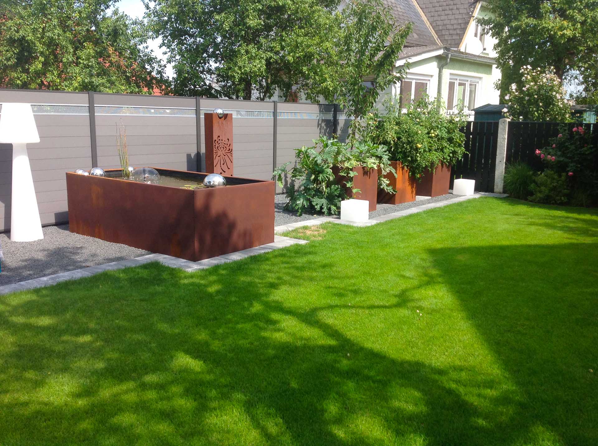 Garten mit grünem Rasen, braunen Pflanzkübeln, Wasserspiel und grauem Sichtschutzzaun bei sonnigem Wetter