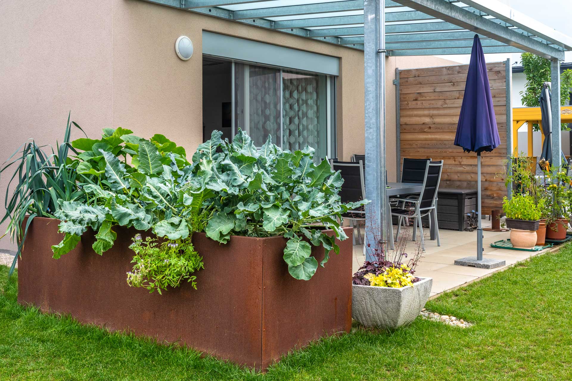 Großes Roststahl-Hochbeet mit verschiedenen Pflanzen vor einer Terrasse mit Glasüberdachung, Gartentisch mit Stühlen und geschlossenen Sonnenschirm