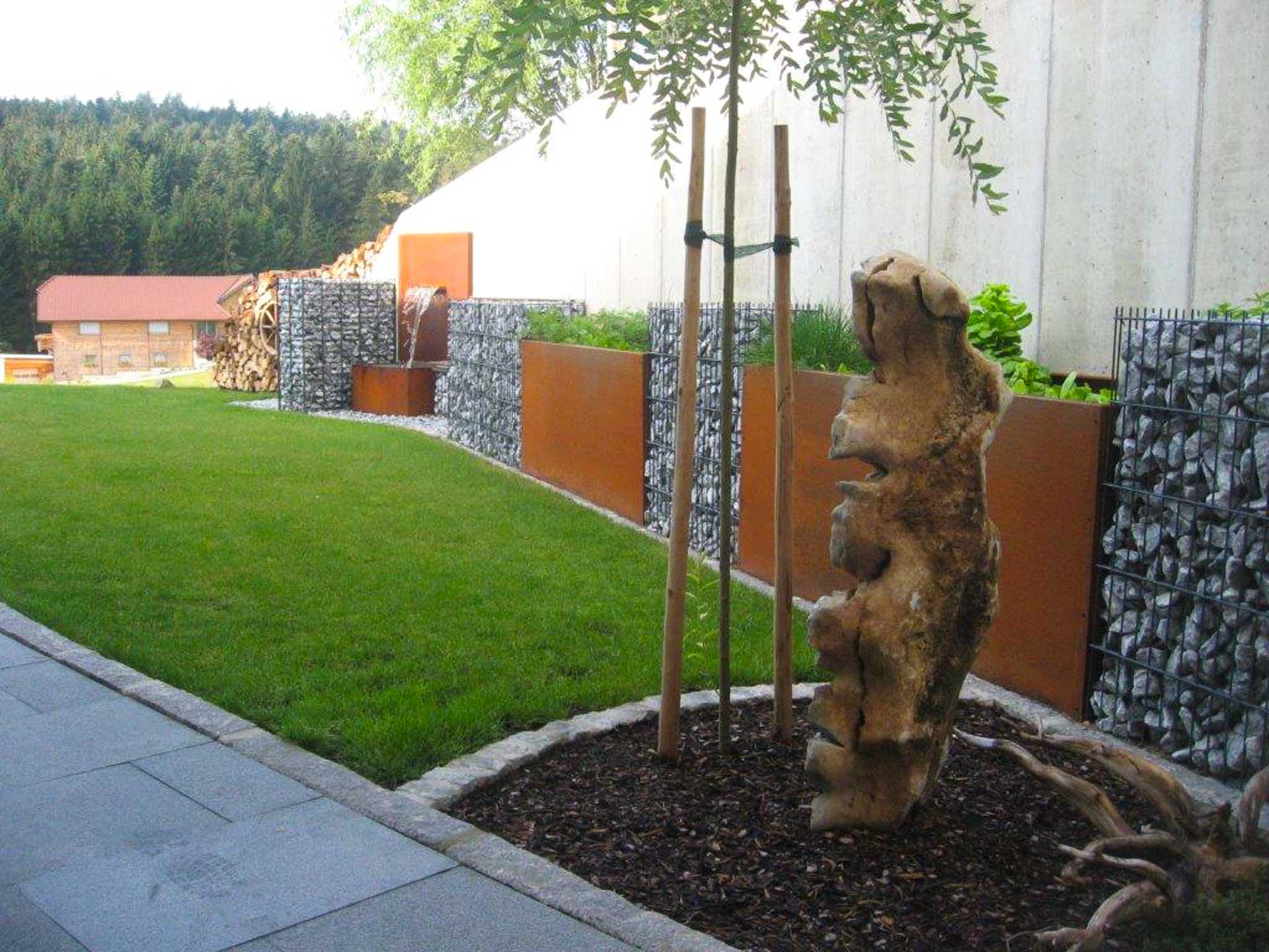 Moderner Gartengarten mit Gabionenwänden, Wasserbrunnen und dekorativem Baumstamm Garten mit grüner Rasenfläche, Gabionenwänden aus Steinkörben mit Cortenstahlelementen, Wasserbrunnen und dekorativem Baumstamm im Vordergrund