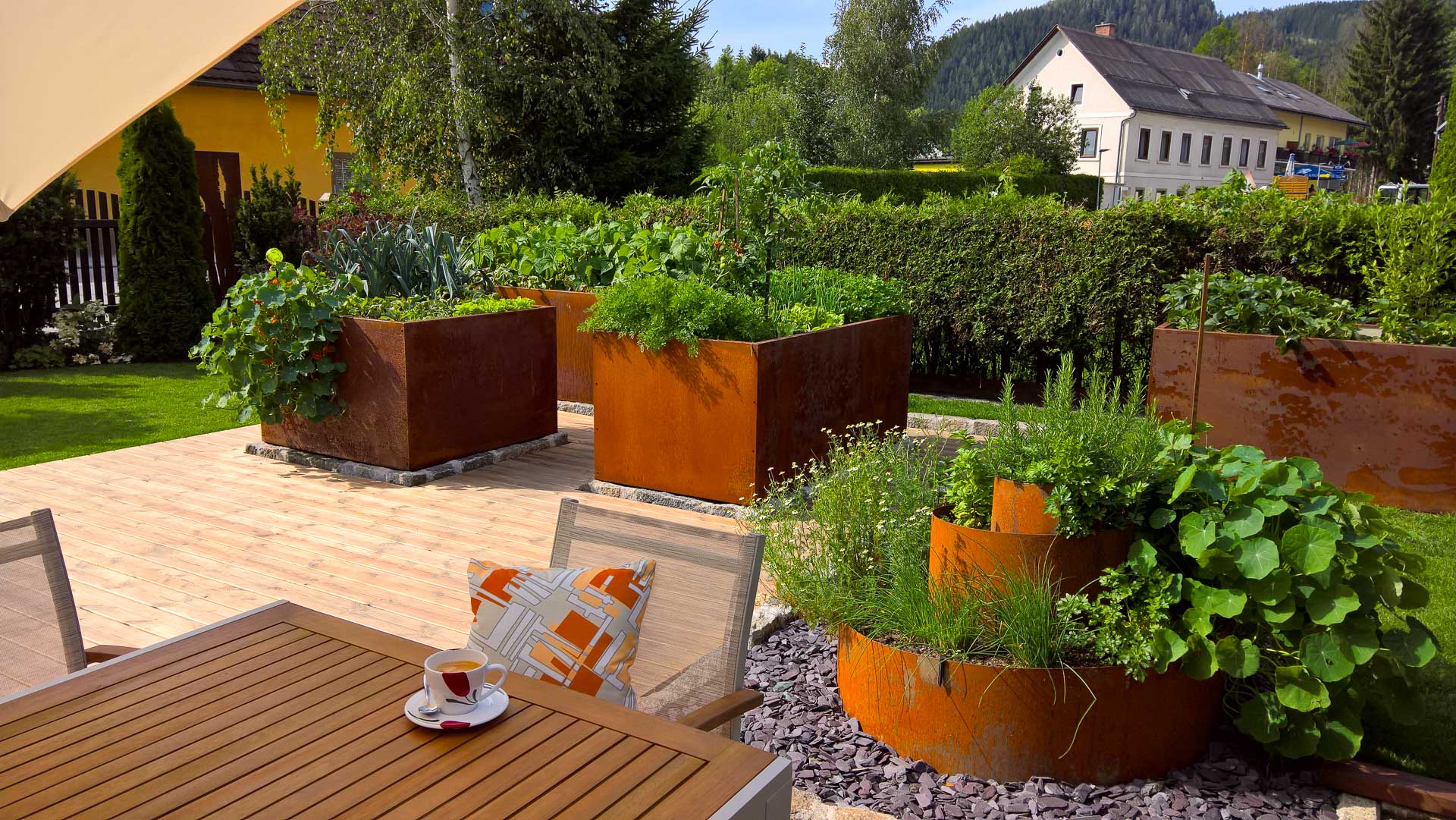 Hochbeete aus Roststahl mit verschiedenen Kräutern und Gemüse auf Holzterrasse im Garten Garten mit mehreren rostbraunen Hochbeeten, bepflanzt mit frischen Kräutern und Gemüse, Holzterrasse mit Stühlen und Tisch, Tasse Kaffee auf dem Tisch