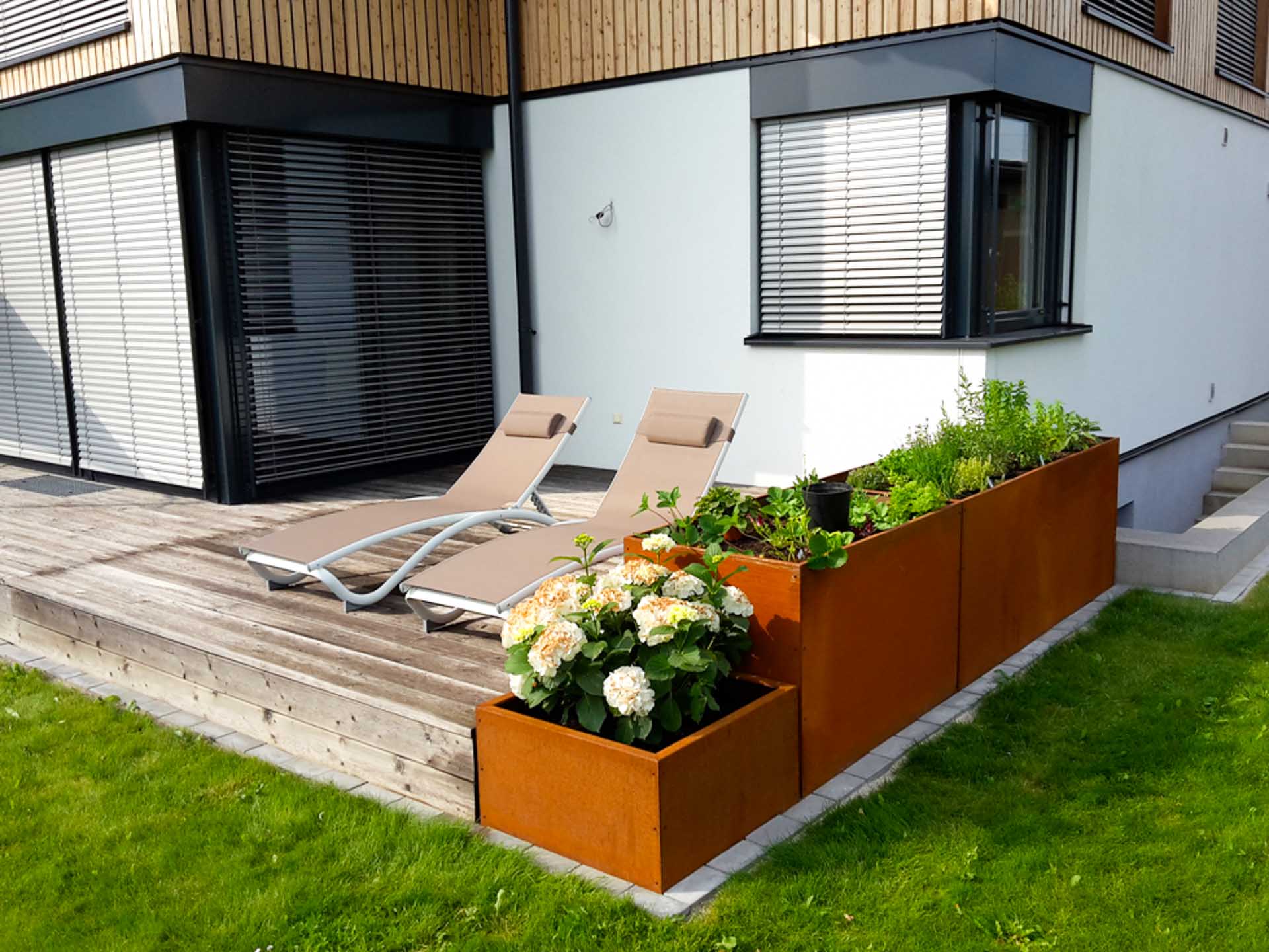 Terrasse mit zwei beigen Sonnenliegen und Hochbeeten aus rostfarbenem Cortenstahl, bepflanzt mit Blumen und Kräutern vor einem modernen Haus mit weißen Wänden und Holzverkleidung